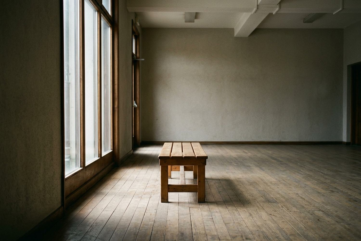 A bare wooden bench by a tall window in a quiet hall, one band of light on the floor.